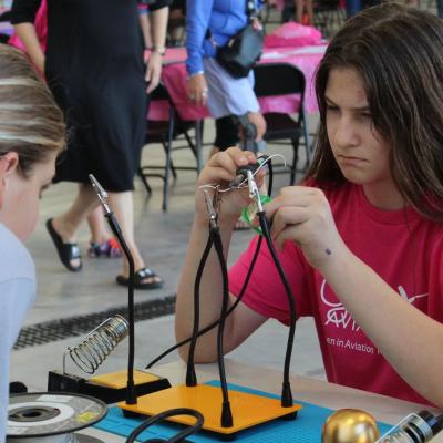 Girl in pink shirt focused on electronics project with tools on table.