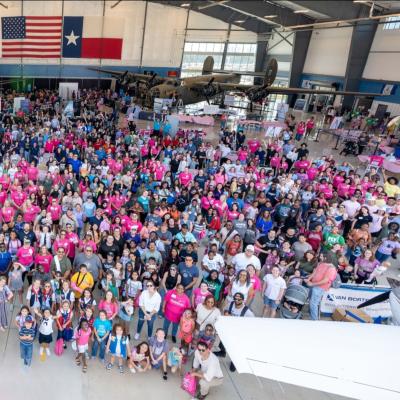 Large group of people inside a hangar, wearing vibrant shirts, under U.S. and Texas flags.