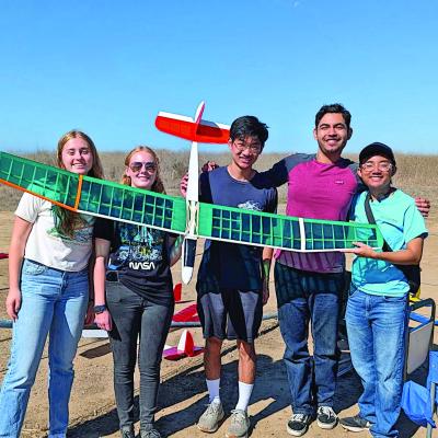 Five people outdoors holding a model plane, smiling under a clear blue sky.