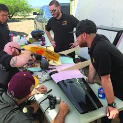 Group of men working on a project outdoors, with tools and a black object on a table.