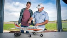 Two men examine a model airplane on a table outdoors.