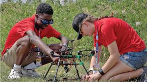Two people in red shirts assembling a small drone outdoors.