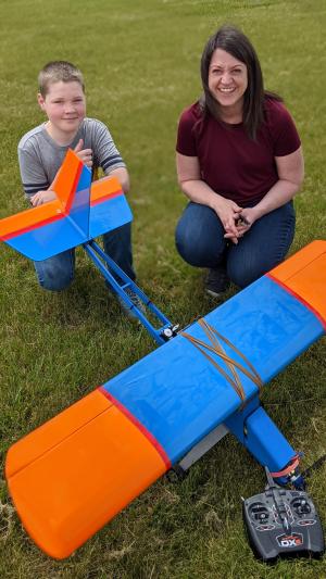 Child and adult smiling beside a colorful model plane on grass.