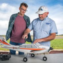 Two men examine a model airplane on a table outdoors.