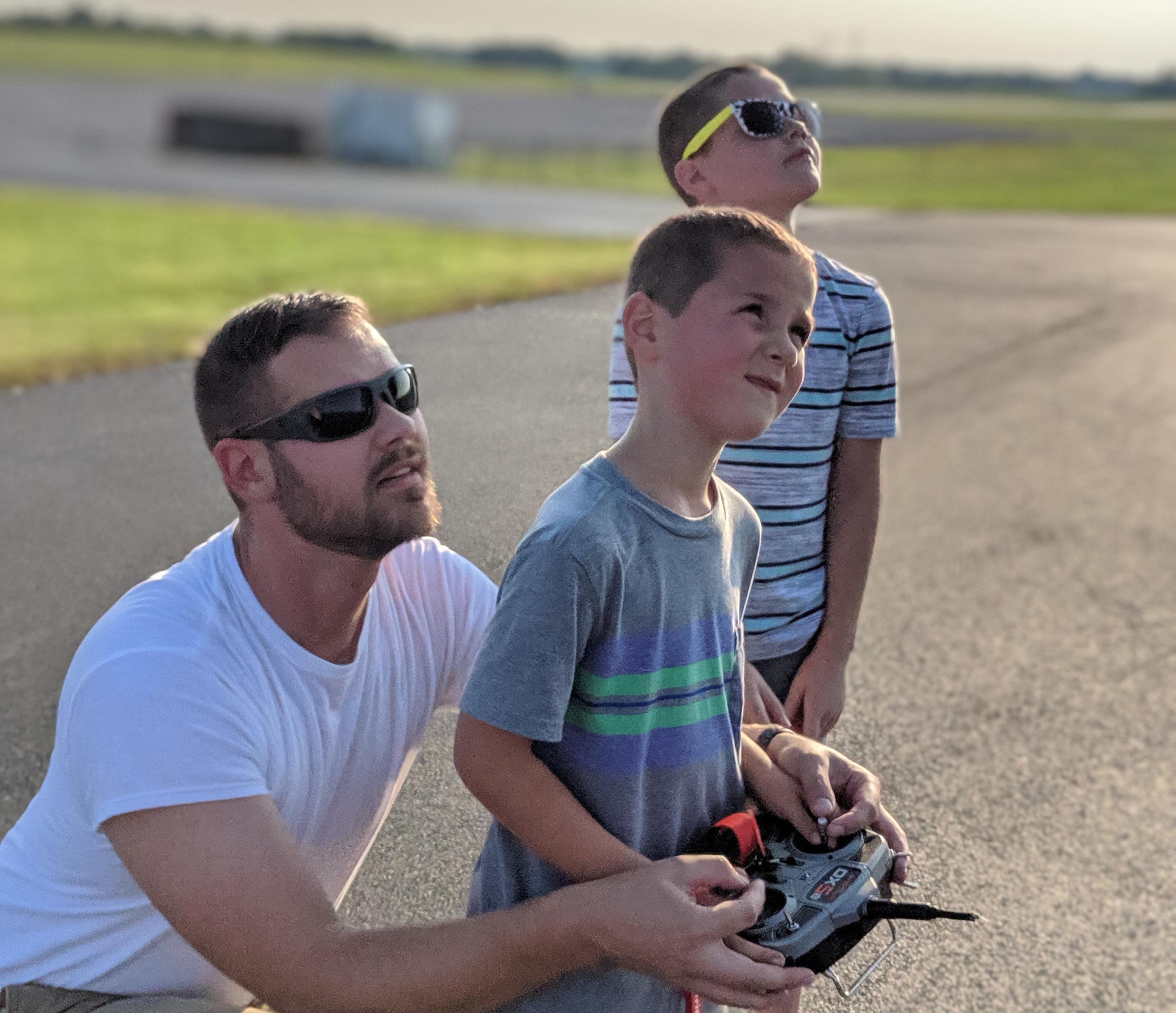 Father and two kids flying a model aircraft, looking skyward on a sunny day.