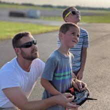 Father and two kids flying a model aircraft, looking skyward on a sunny day.