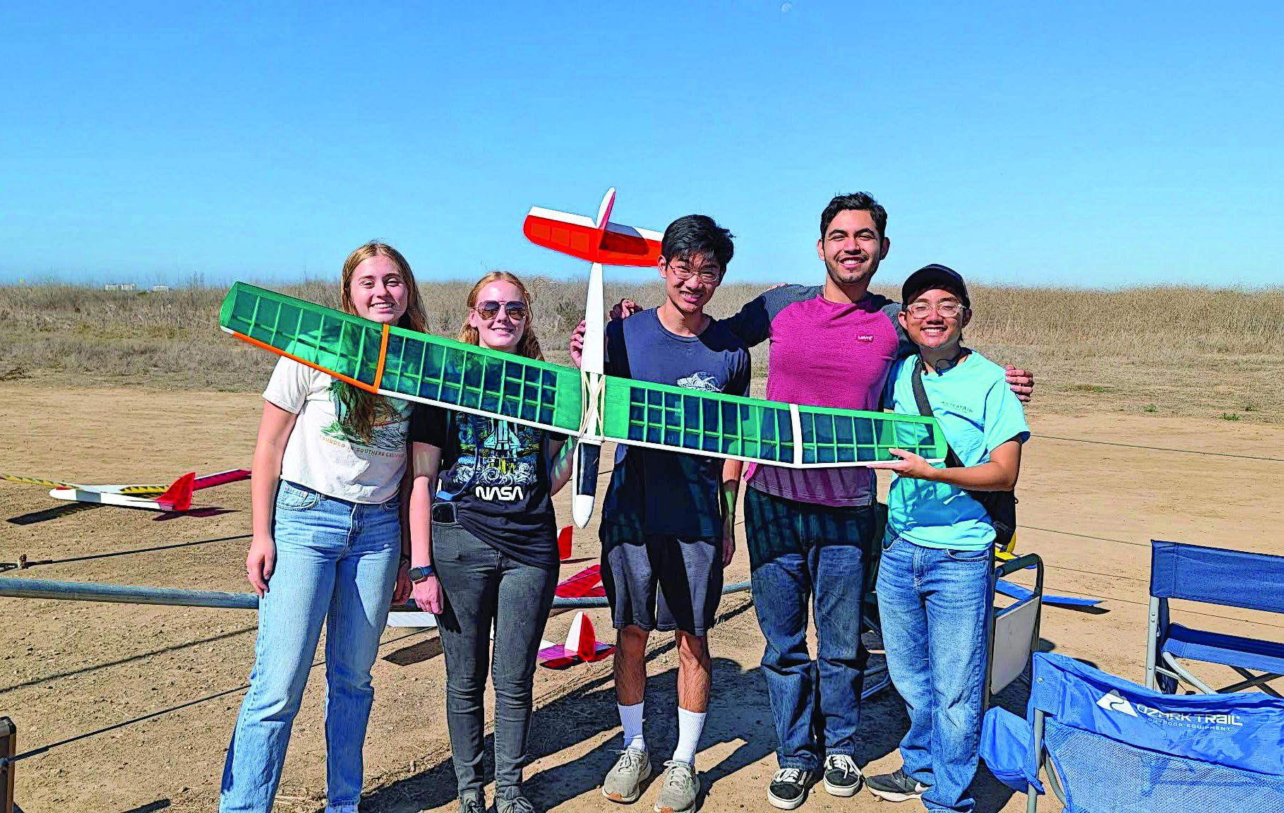 Five people outdoors holding a model plane, smiling under a clear blue sky.